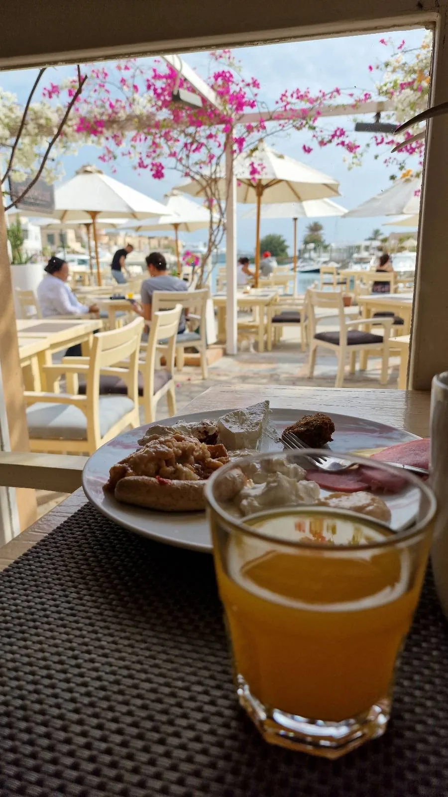 A close-up of a table with a glass of orange juice and a plate of food. The background shows an outdoor seating area with people at tables under umbrellas and blooming pink flowers.
