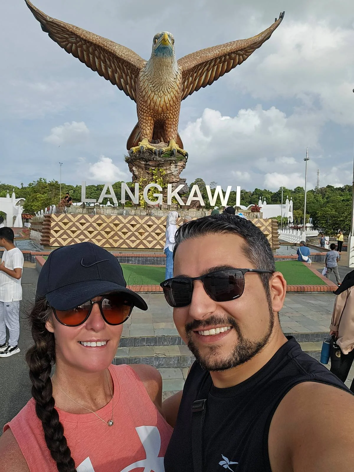 A smiling couple takes a selfie in front of the large eagle statue at Dataran Lang in Langkawi, Malaysia, with the word "LANGKAWI" displayed behind them.