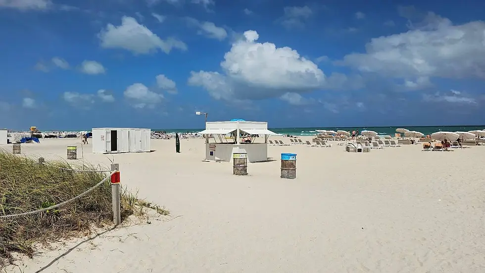 Sandy beach with empty chairs and umbrellas under a blue sky. A white booth and bins labeled "Recycling" and "Trash" in the foreground.