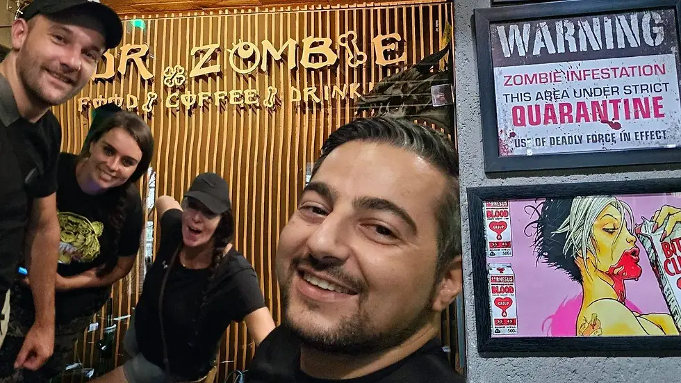 Four people smiling in front of "Dr. Zombie" café sign. Wall displays "Warning: Zombie Infestation" and colorful zombie art. Mood is playful.