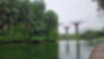Lush green trees line a pond with futuristic tree-like structures in the background. Overcast sky and reflections on the water create a serene mood.