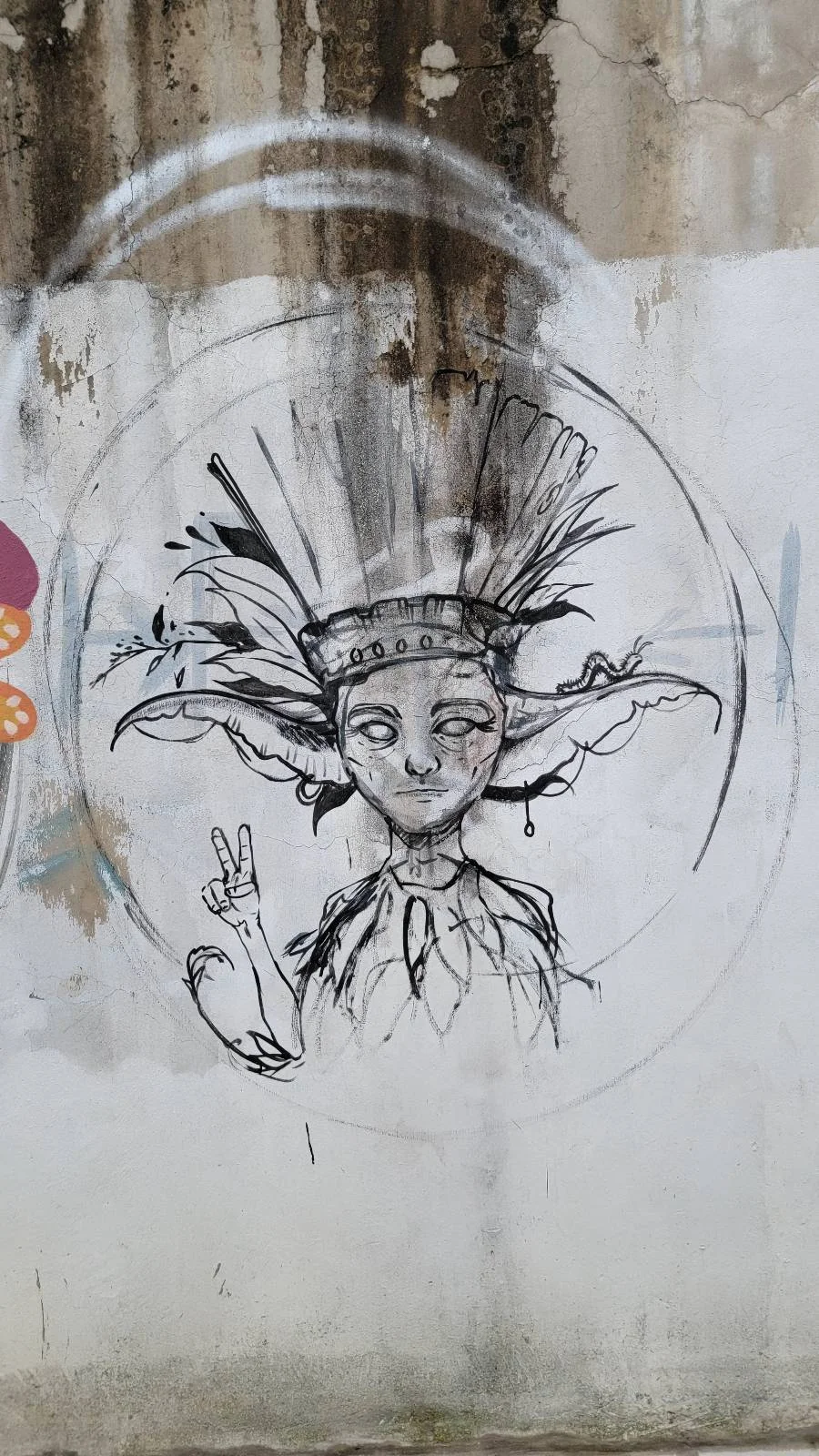 A black and white graffiti piece depicts a character with a tall, feathery headdress and exaggerated facial features. The character is making a peace sign with one hand. The art is on a weathered concrete wall.
