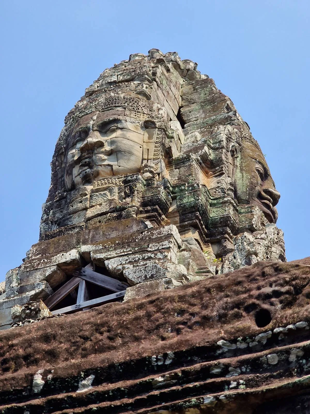 A close-up view of a stone tower at Bayon Temple in Cambodia, featuring large, intricately carved faces on its surface against a clear blue sky.