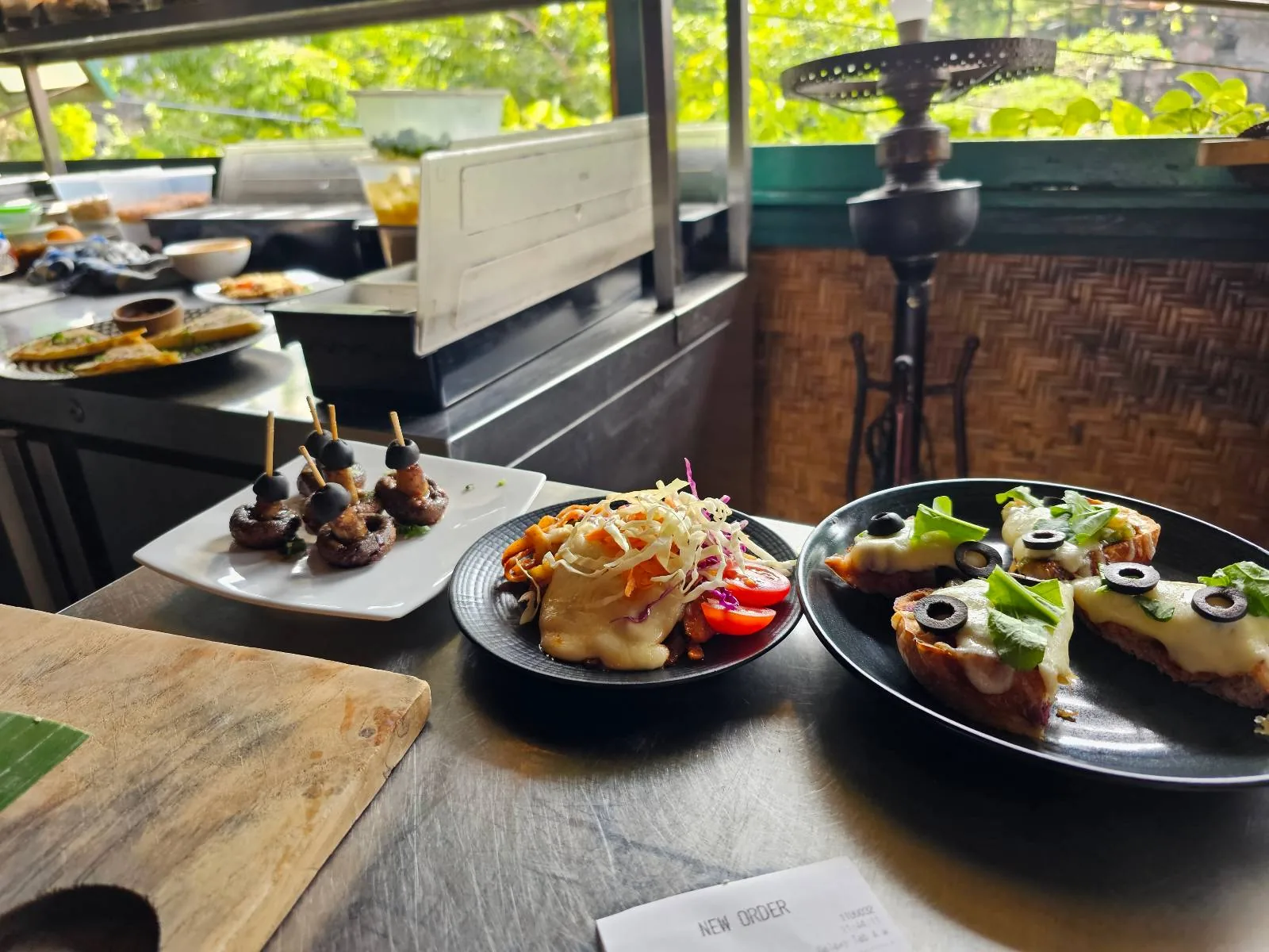 Small plates of assorted appetizers, including skewered items, a salad with shredded cheese and cherry tomatoes, and slices of bread topped with greens and olives, are arranged on a table by a window with greenery outside.