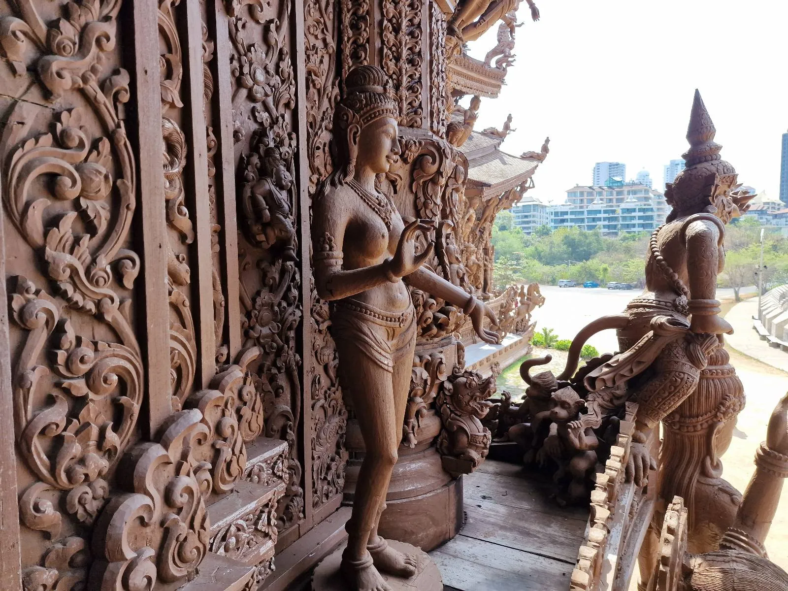 A detailed wooden sculpture of a mythical figure is set against intricately carved panels. Another similar sculpture stands in the background. The setting overlooks a lush, green landscape and distant cityscape under a clear sky.