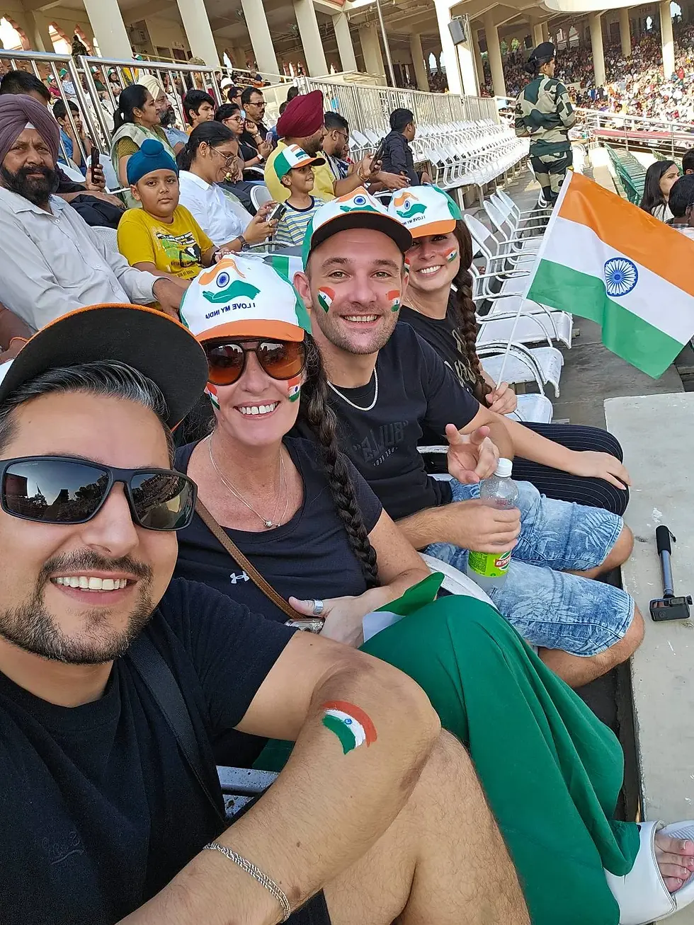 Group of people with Indian flag face paint and caps, smiling at a stadium. Crowd in background, one holding an Indian flag. Celebratory mood.