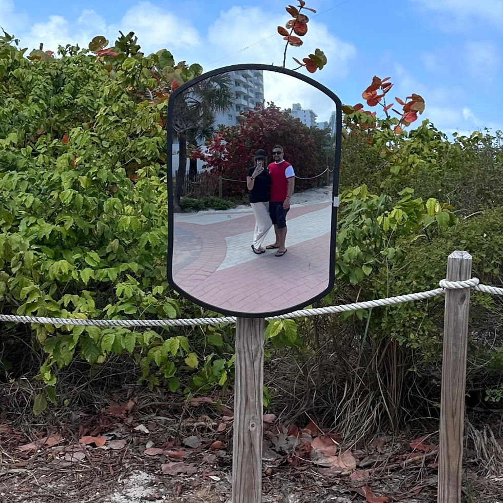 Couple reflected in a convex mirror on a leafy path. They stand close, smiling. Mirror is framed by greenery and a wood post with rope.