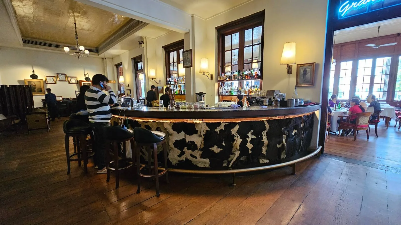 A bar with a marble counter and high stools in a spacious room. A person sits at the bar, and a few people are visible in the background through the doorway. The room features wooden floors, tall windows, and a neon sign.