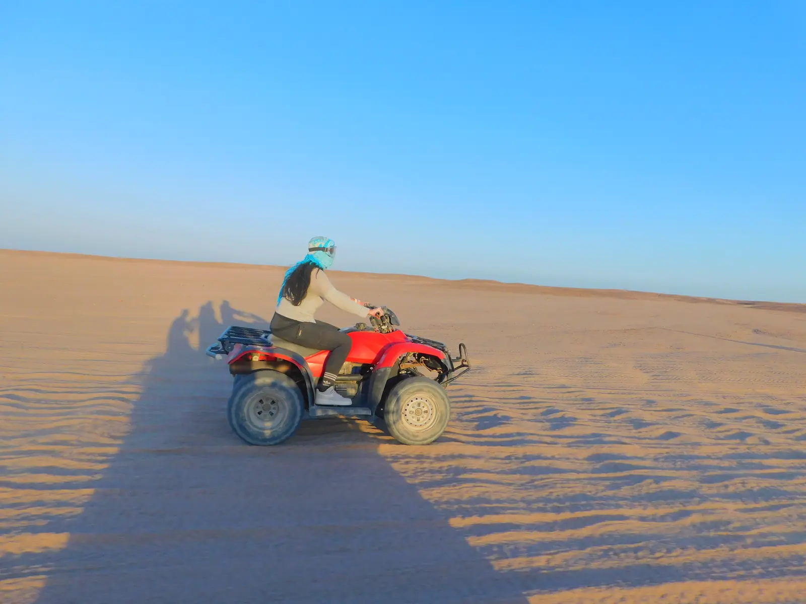 A person wearing a helmet is riding a red ATV across a vast, sandy desert under a clear blue sky. Long shadows are cast on the sand, and the terrain is flat and expansive.