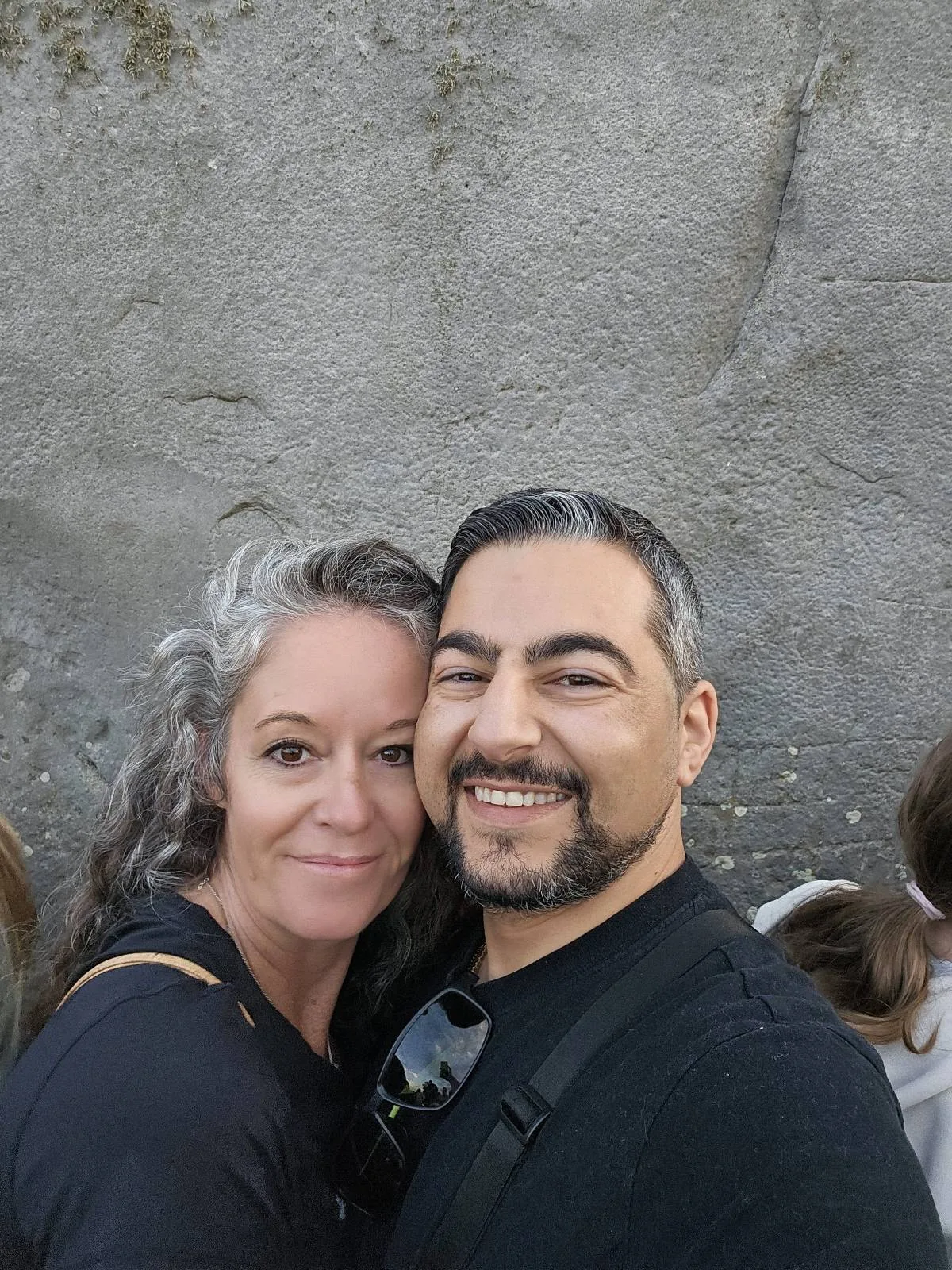 A smiling couple stands close together in front of a textured gray wall, posing for a selfie. The woman has gray-streaked hair and is wearing a black top; the man has dark hair and a beard.