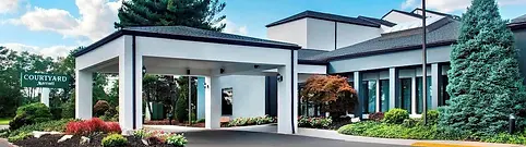 Front view of a Courtyard by Marriott hotel featuring a modern white and gray exterior with a covered entrance. The landscape includes manicured shrubs, green grass, and a clear, blue sky with fluffy clouds overhead.