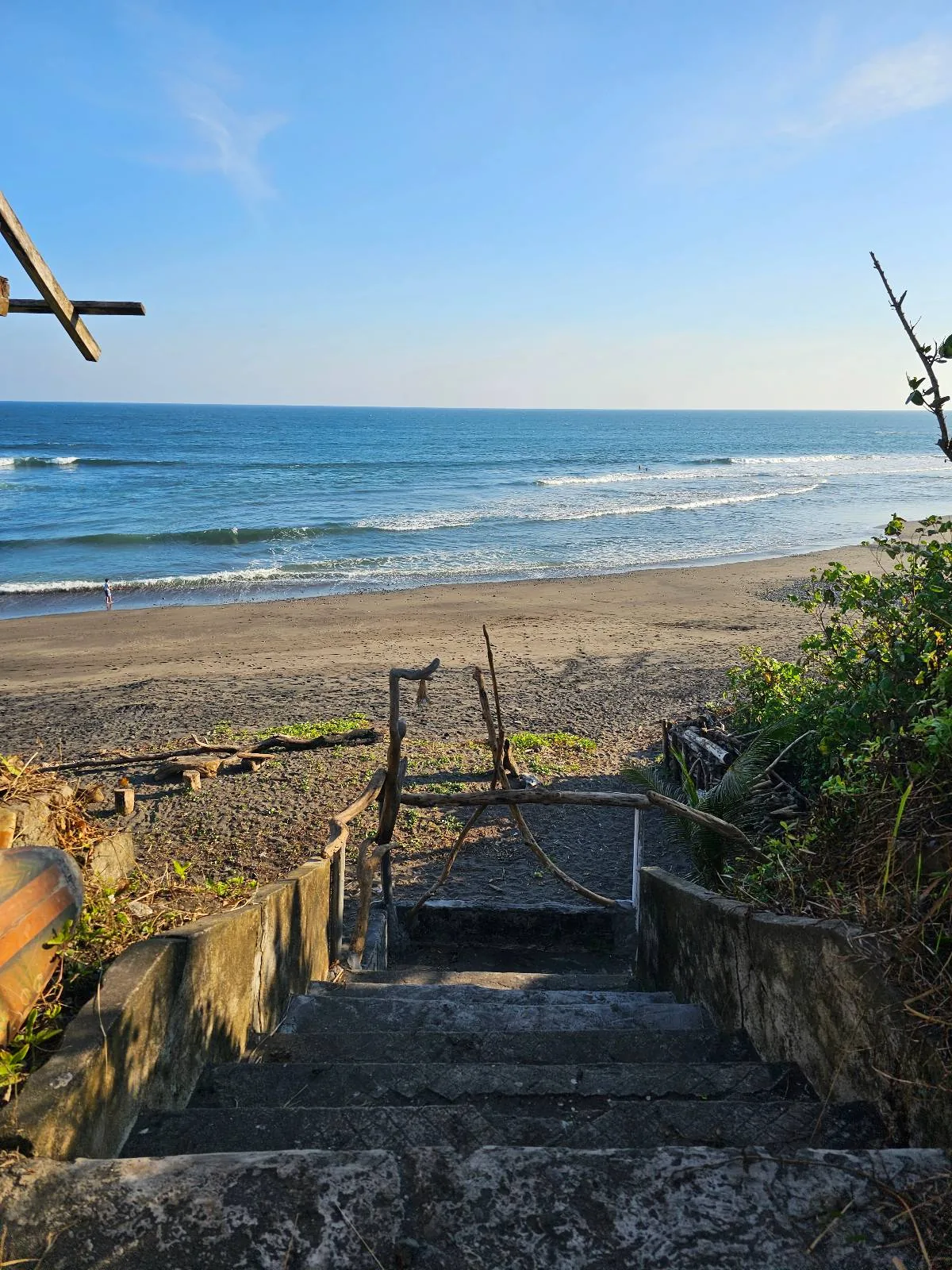 A stone staircase leads down to a sandy beach with gentle waves under a clear blue sky. Sparse vegetation flanks the stairs, and the ocean stretches into the horizon, creating a serene coastal scene.