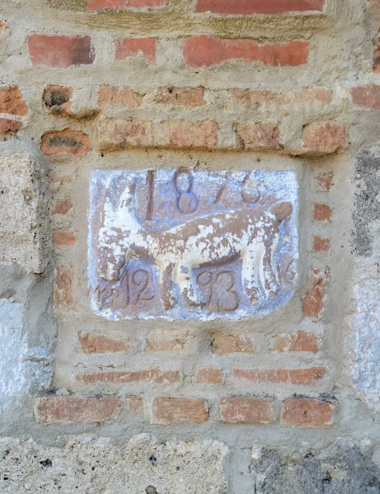 A weathered stone plaque mounted on a brick and stone wall, engraved with the year "1876" at the top and "1293" at the bottom, with some of the numbers worn and faded.