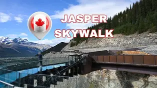 Person on glass walkway at Jasper Skywalk, surrounded by mountains and trees. Clear sky with "Jasper Skywalk" text overlay and Canadian flag.