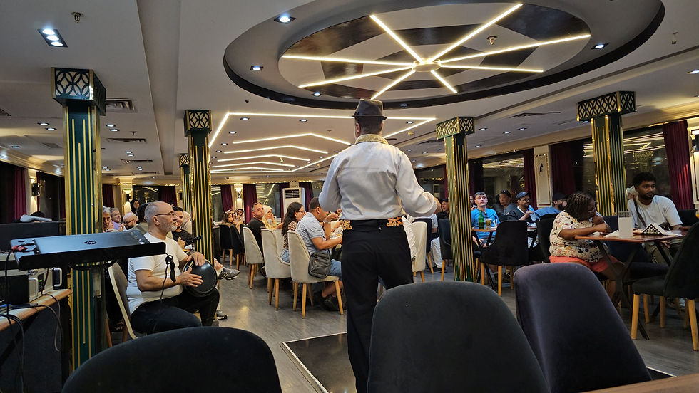 Man in hat performing for seated audience in a stylish venue with circular ceiling lights and green pillars. Some attendees are dining.