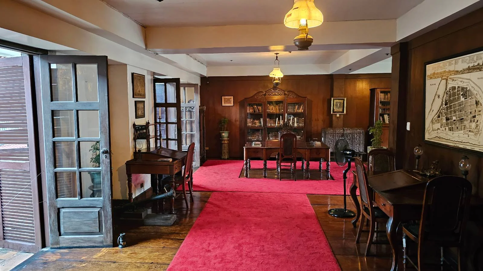 A sophisticated room with wooden floors and paneling features a red carpet leading to shelves of books. The area is furnished with dark wood tables and chairs, lit by warm hanging lamps. A large drawing is displayed on the right wall.