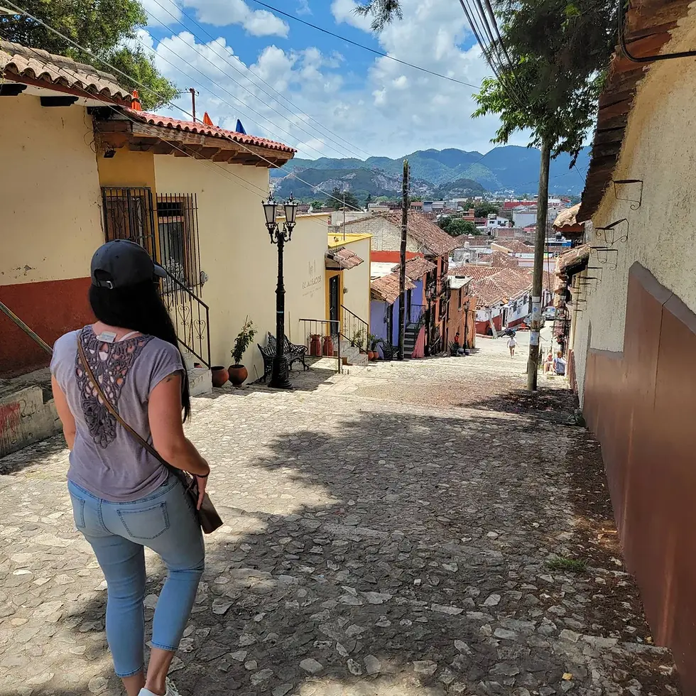 Woman walks down a cobblestone street in a charming town. Colorful houses, mountainous backdrop, sunny day. Relaxed, scenic atmosphere.