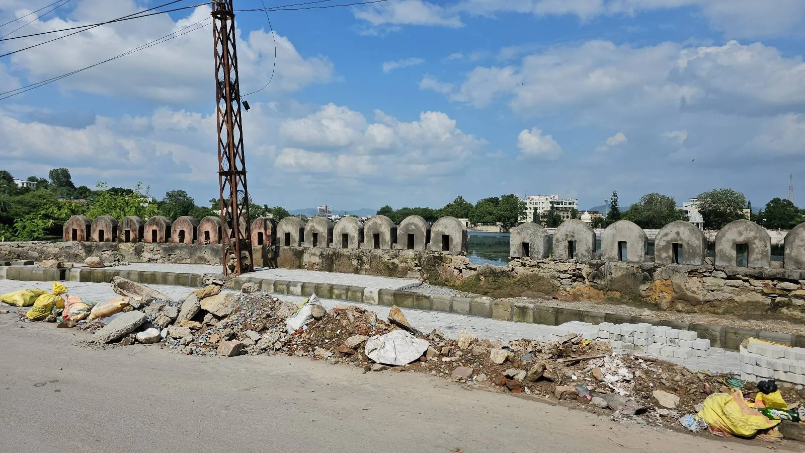A row of old, arched stone structures lines a waterway behind a construction site with scattered debris and yellow bags under a partly cloudy sky.