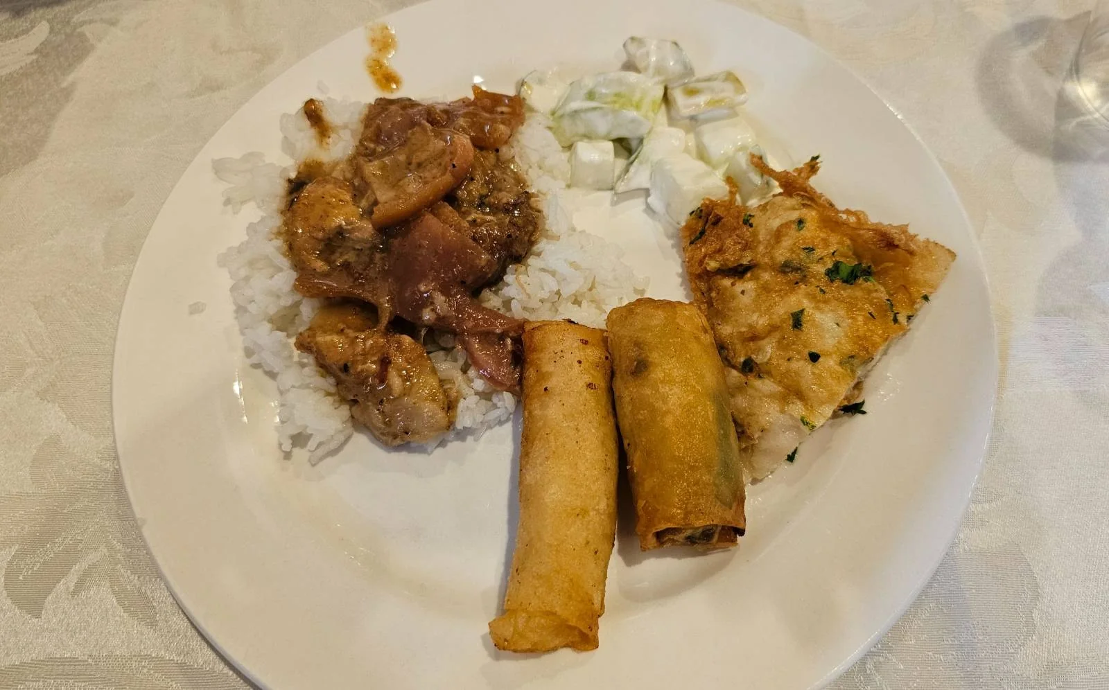 A plate with rice, meat stew, cucumber salad, a spring roll, and a piece of omelette on a white tablecloth.