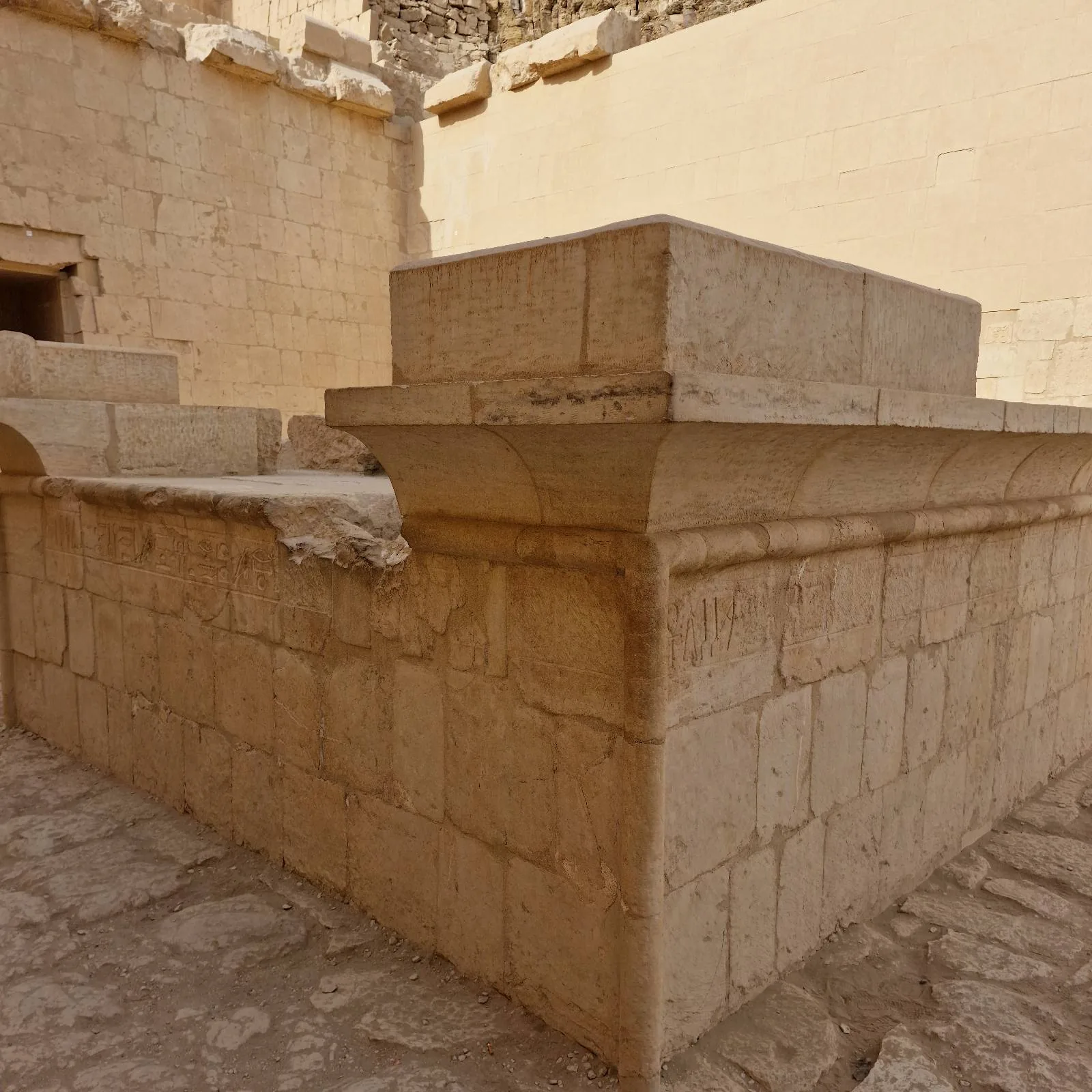 A stone structure with rectangular walls, possibly an ancient tomb or altar, stands in a sunlit, open area with beige stone flooring and high walls in the background.