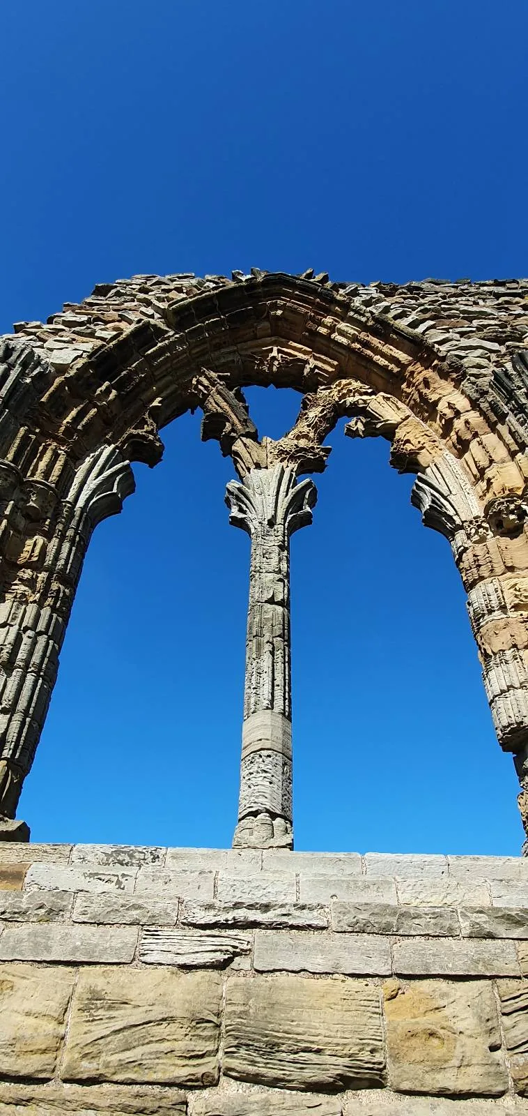 An ancient stone window with intricate carvings, set against a clear blue sky. The weathered structure stands prominently, showcasing historical architecture.