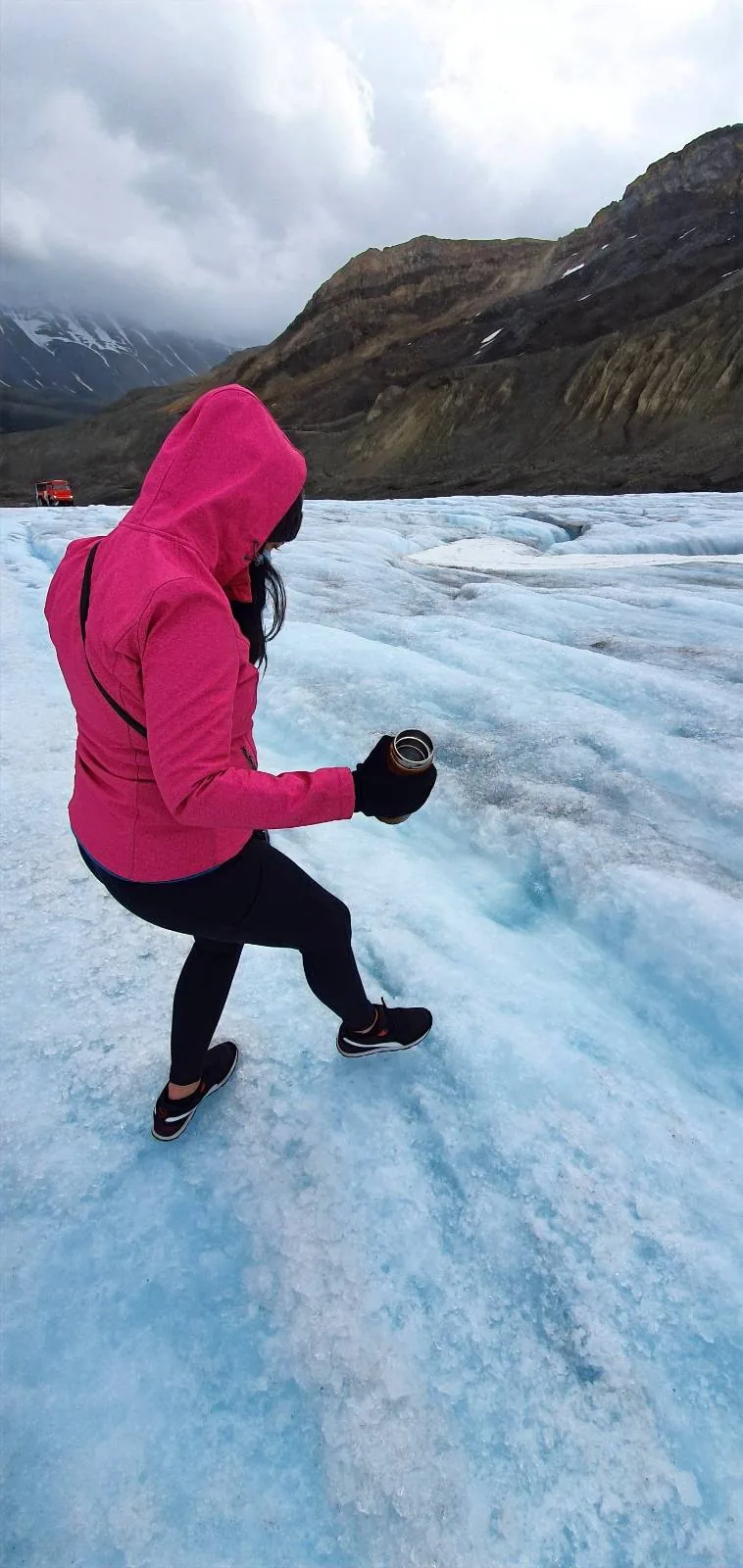 A person in a bright pink jacket and gloves walks carefully on a blue glacier, with mountains and cloudy skies in the background.