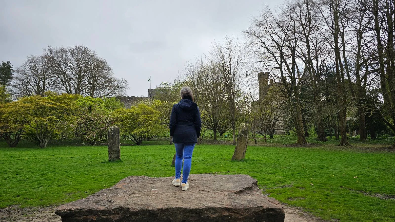 Person standing on a large stone, looking at a distant castle in a lush green park.