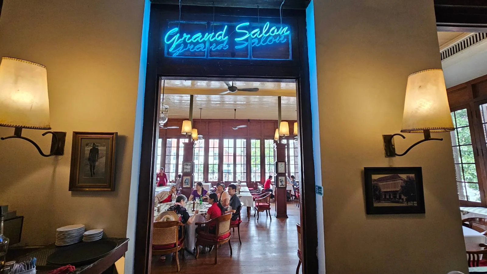 Entrance to a restaurant with a neon sign reading "Grand Salon." Inside, diners sit at tables under warm lighting. Walls are adorned with framed pictures, and large windows offer a view of greenery outside.