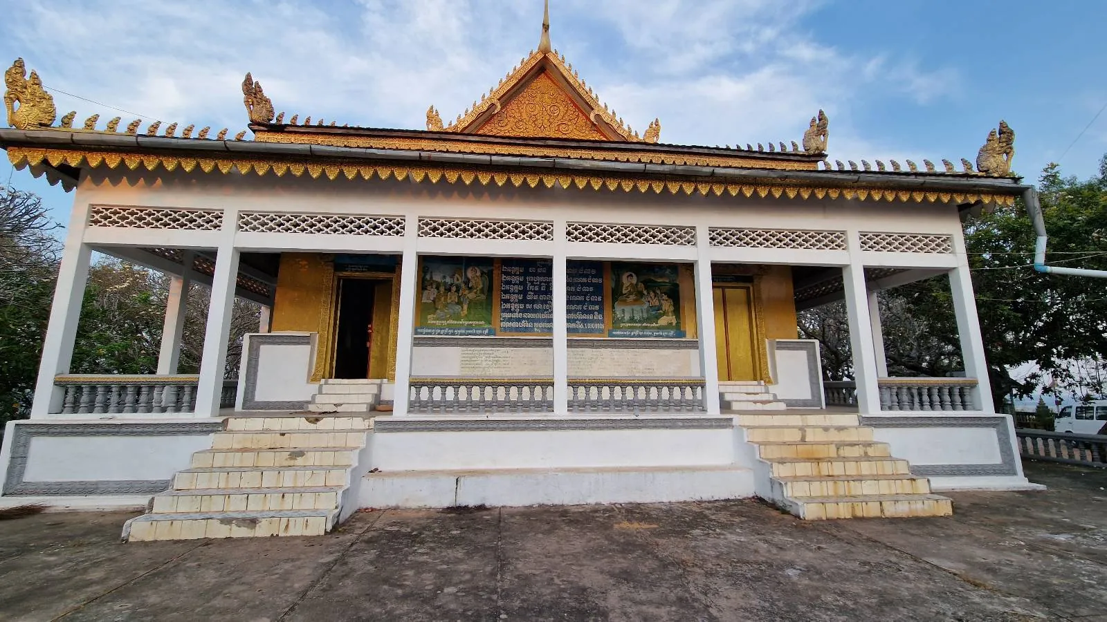 A decorative gate with golden details and script in a foreign language stands at the top of stone steps, surrounded by trees. A person is seen climbing the stairs toward the gate.