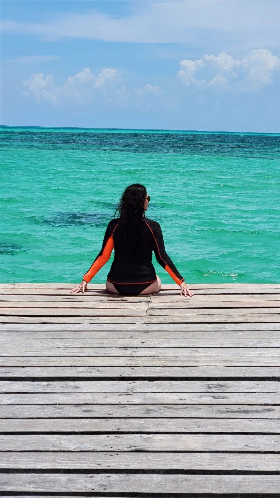 A person with long hair sits on a wooden deck, facing a turquoise ocean under a partly cloudy sky. They are wearing a black and orange swimsuit and are looking at the horizon.
