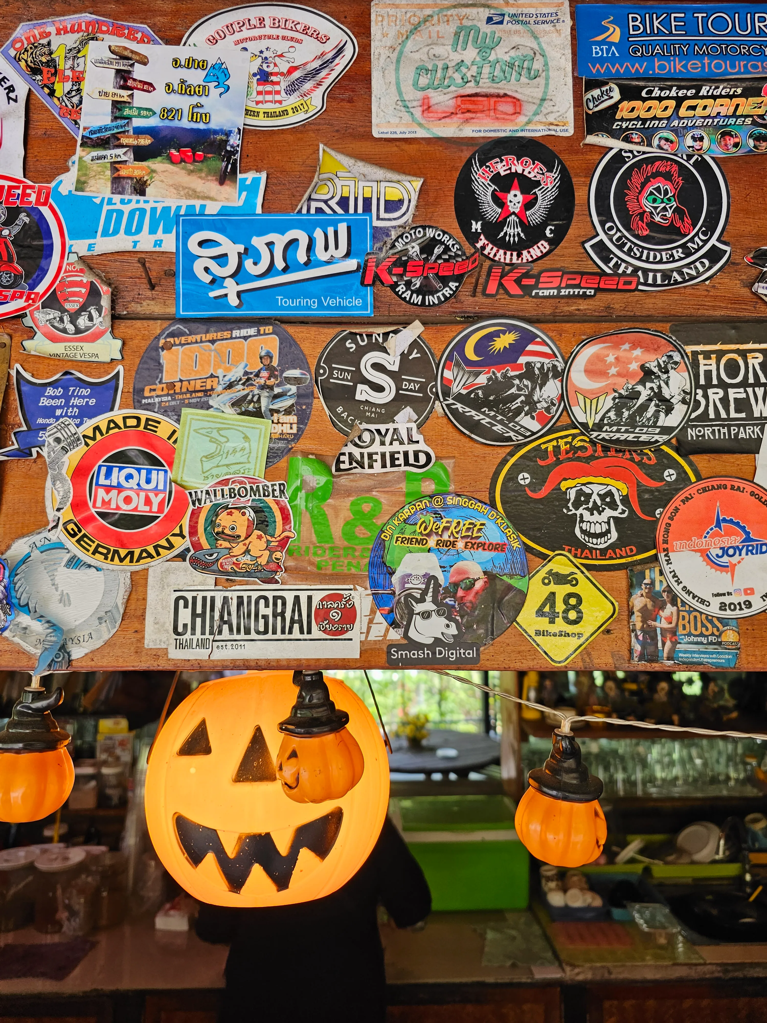 A wooden wall covered with colorful stickers and logos, with two orange jack-o’-lantern decorations and a person holding a large pumpkin mask in front of their face at the bottom of the image.