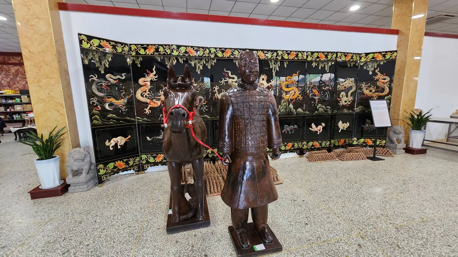 Two bronze statues stand in front of an ornate folding screen decorated with dragons in a brightly lit room with tiled floors. Potted plants and carved stone figures are placed along the sides.