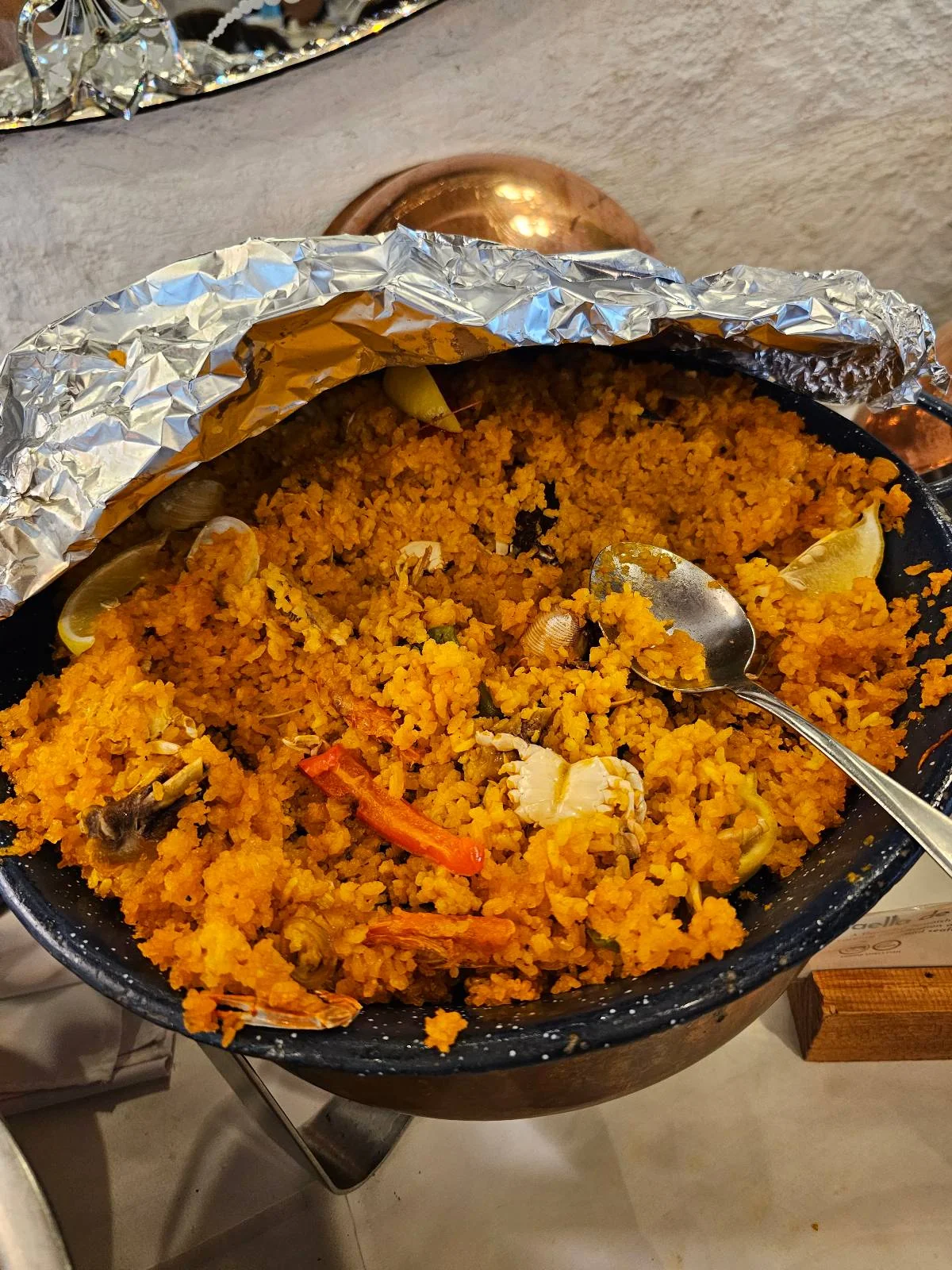 A large dish of yellow rice mixed with vegetables and pieces of meat, partially covered with aluminum foil, with a serving spoon placed on top.