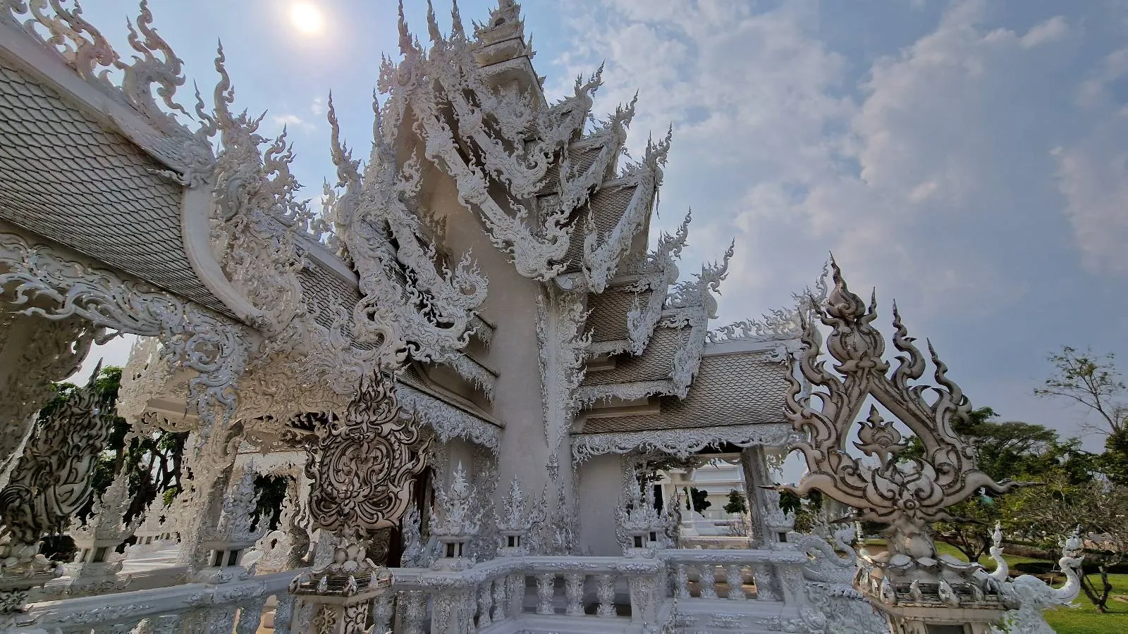 Ornate white temple with intricate carvings and spires, set against a partly cloudy sky. Sunlight highlights the detailed architecture, and greenery is visible in the background.