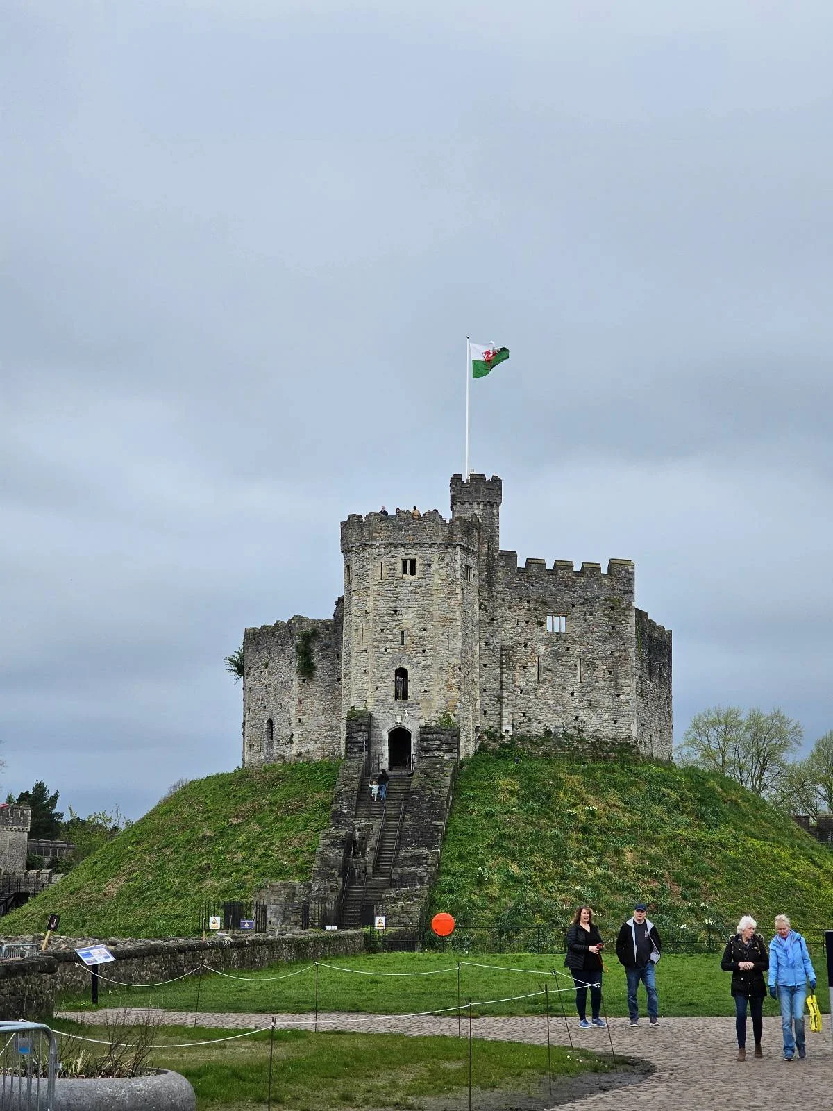 Stone castle on a grassy hill with an irish flag waving on top, under a cloudy sky, with a few visitors at the base.