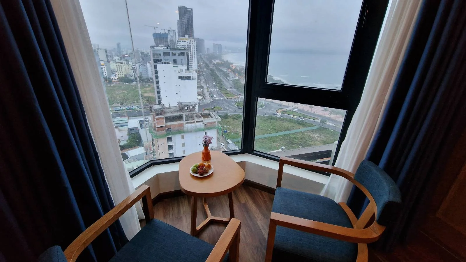 A cozy corner with two chairs and a small round table by a large window, overlooking a cityscape with tall buildings and a distant view of the ocean. The table has a bottle, glasses, and a bowl of fruit. Curtains frame the window.