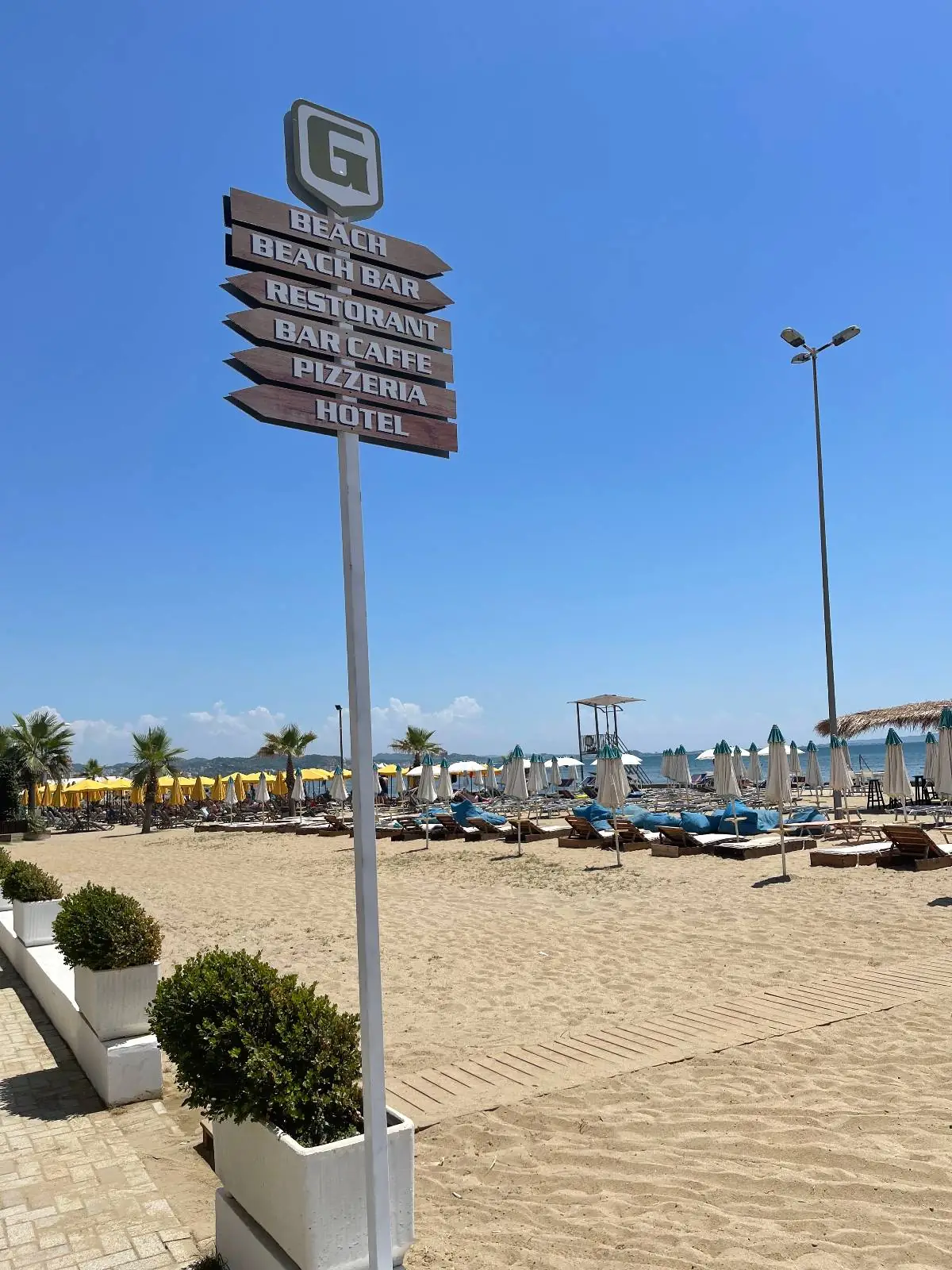 A signpost stands on a sandy beach near potted plants, with directions to various amenities. Sun loungers, umbrellas, and a clear blue sky are visible in the background, creating a relaxing beach scene.