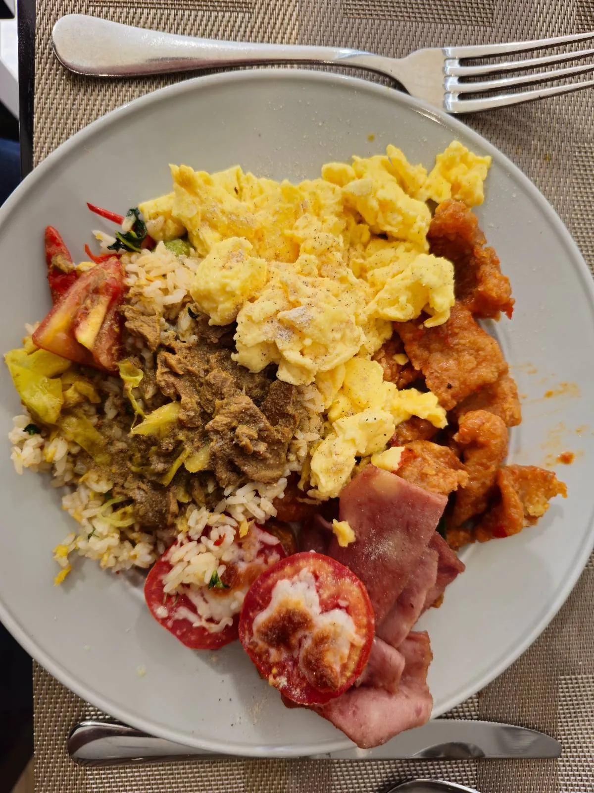 A plate with scrambled eggs, fried chicken pieces, white rice, shredded meat, sliced tomatoes, and pieces of ham, set on a light-colored placemat with a fork and knife beside it.