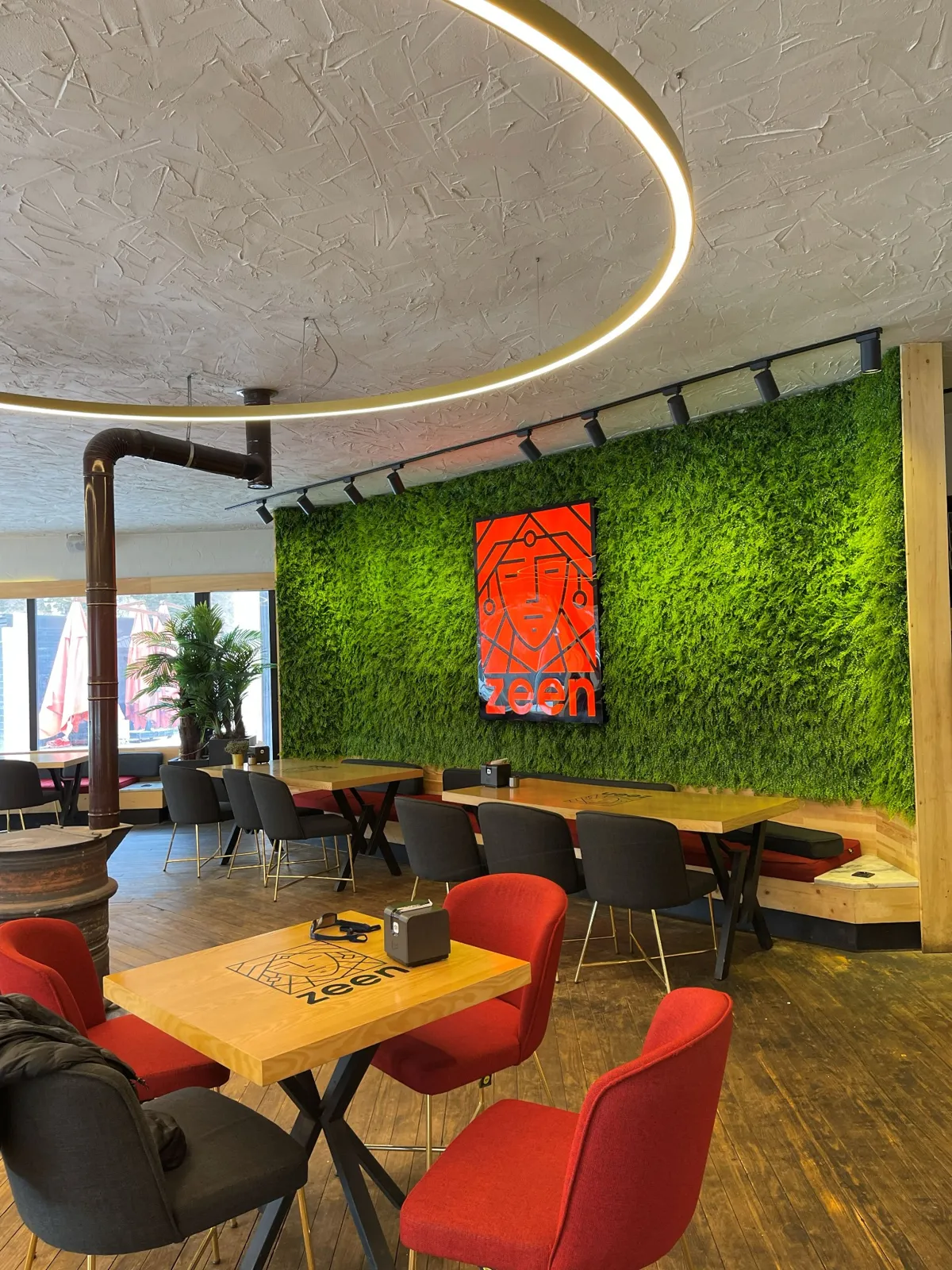 Modern cafe with green moss wall, circular ceiling light, and tables with red and black chairs. A red geometric art piece is displayed on the wall.