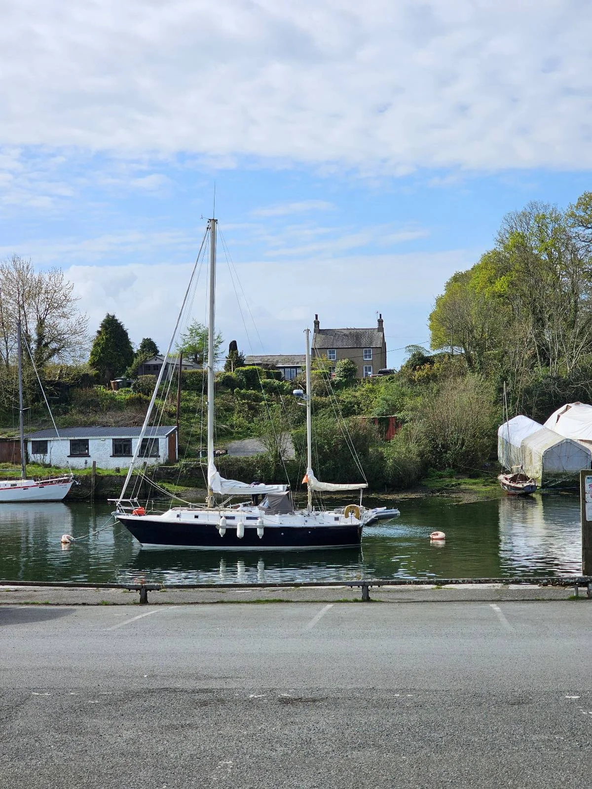 A small sailboat is docked on a calm river near a tree-lined shore, with white buildings and a church visible in the background under a partly cloudy sky.