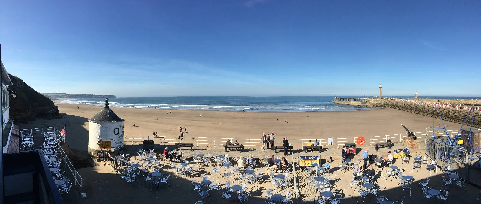 Panoramic view of a beach with a clear blue sky. The sandy shore has numerous white chairs and umbrellas. A small round building with a conical roof is on the left, and a lighthouse stands on a distant pier to the right.