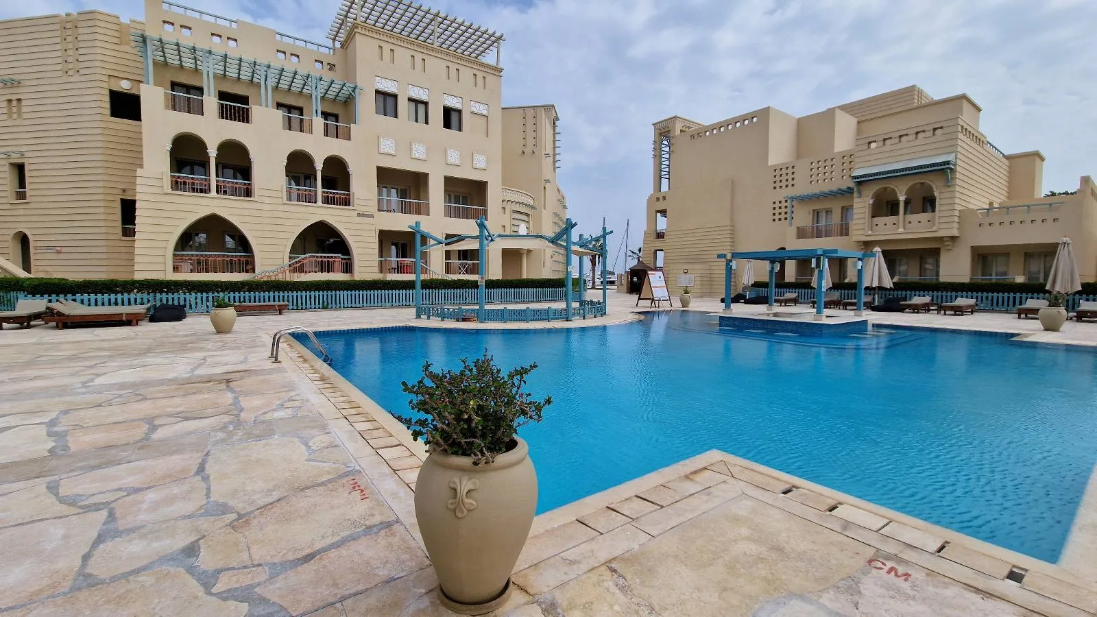Outdoor swimming pool surrounded by beige buildings with balconies and blue pergolas, large potted plants, lounge chairs, and a partly cloudy sky above.