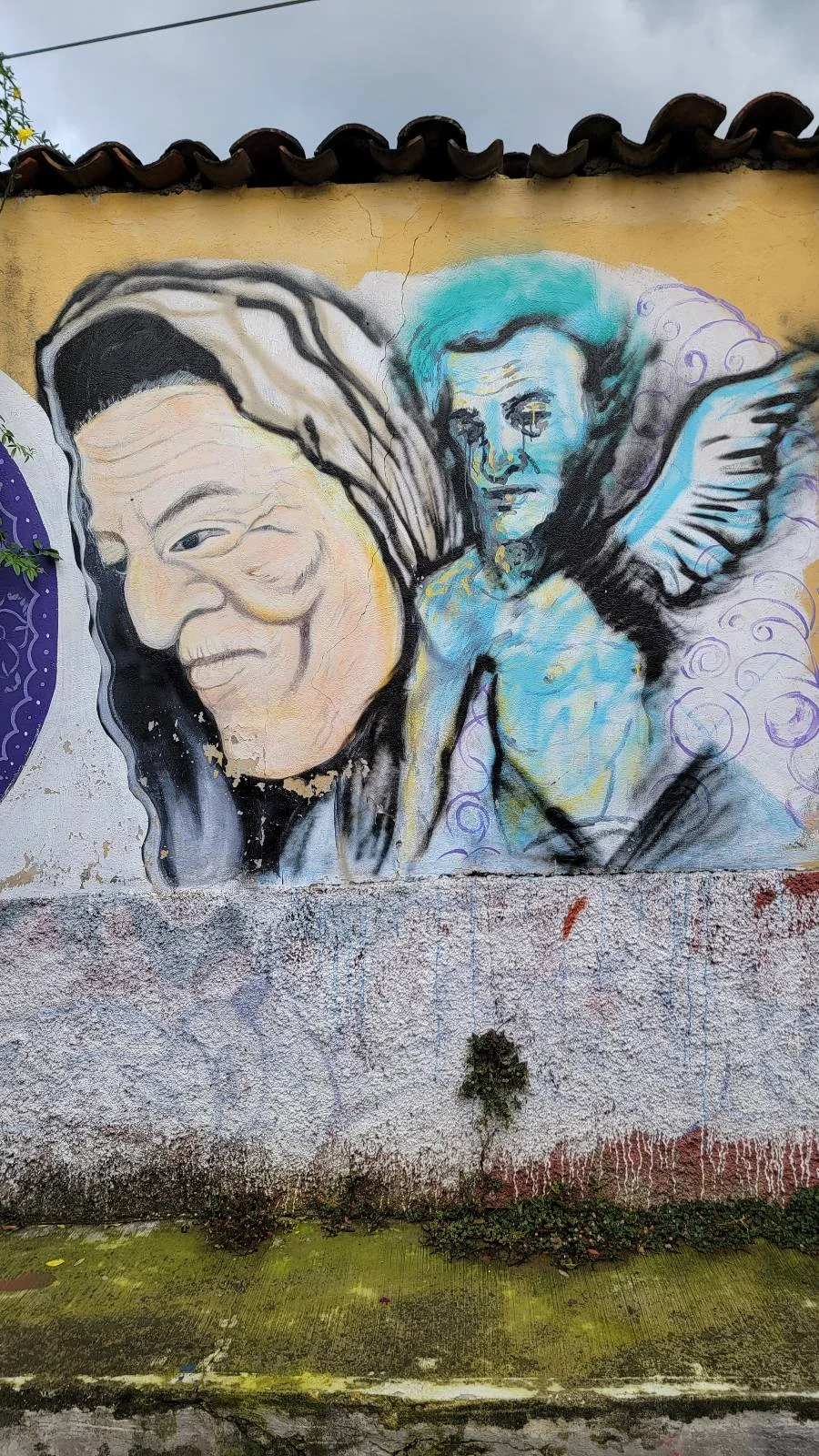 A mural depicting a large face of a person with long hair and an angelic figure with wings beside them. The background features abstract patterns in purple and white on a weathered wall.