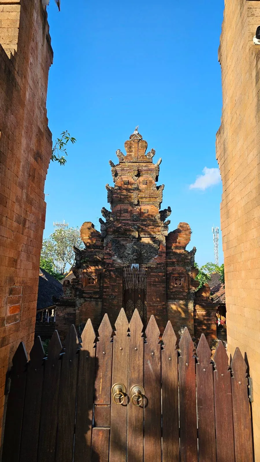 A view through a wooden gate of a traditional Balinese temple entrance, featuring intricate stone carvings. The temple is set against a clear blue sky, framed by two stone structures on either side.