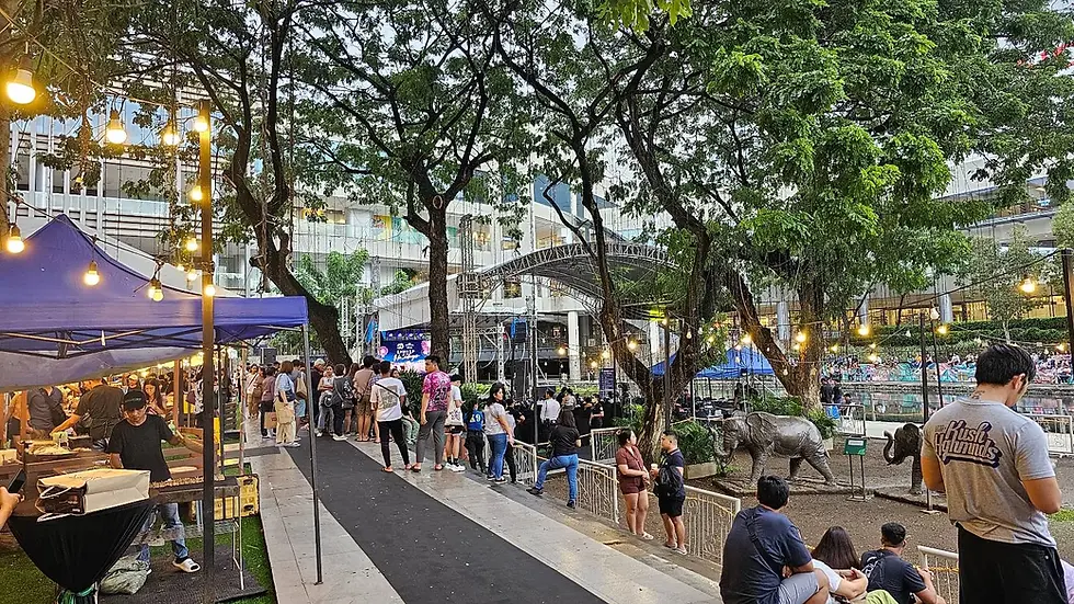 People gather at an outdoor market with food stalls and string lights. A stage is visible in the background, suggesting an upcoming event.