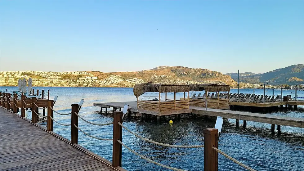 Wooden piers with lounge chairs extend into a calm body of water, with hills and a town visible in the background under a clear blue sky.