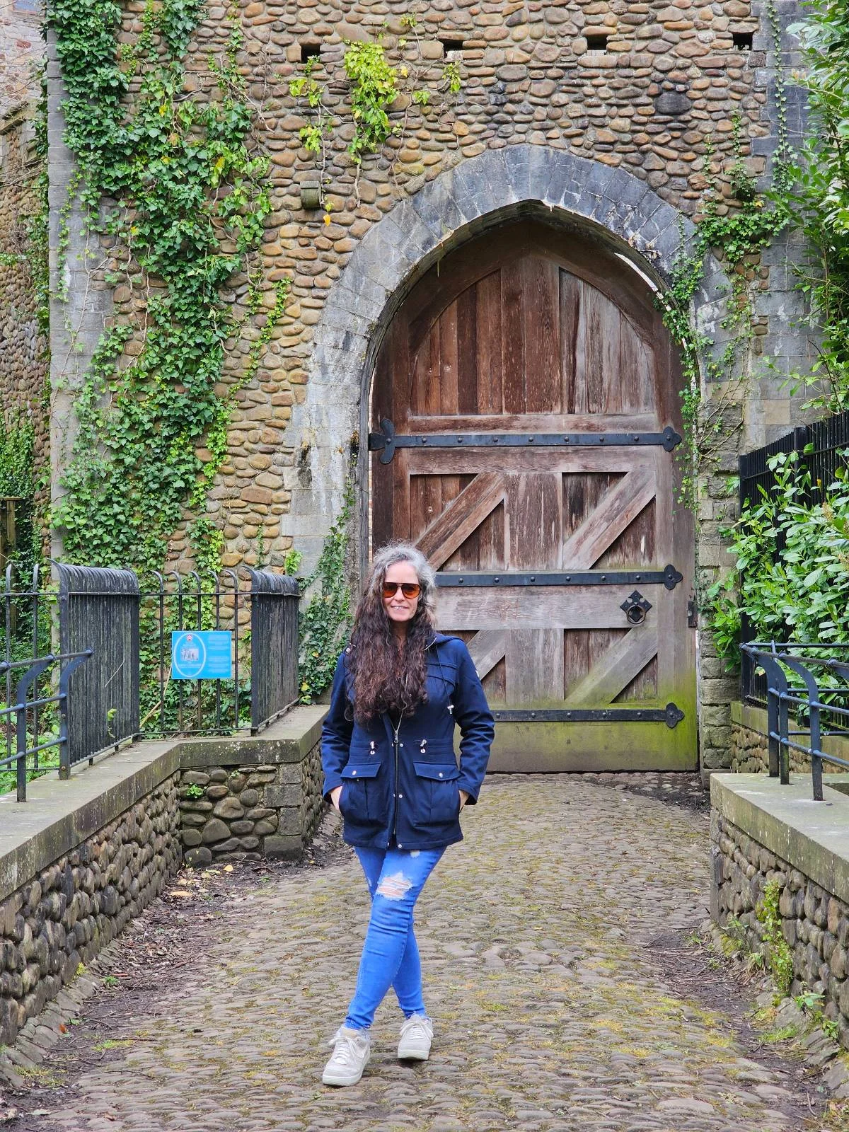 A woman wearing sunglasses, a blue jacket, and jeans standing in front of a large, aged wooden door set in a brick archway.