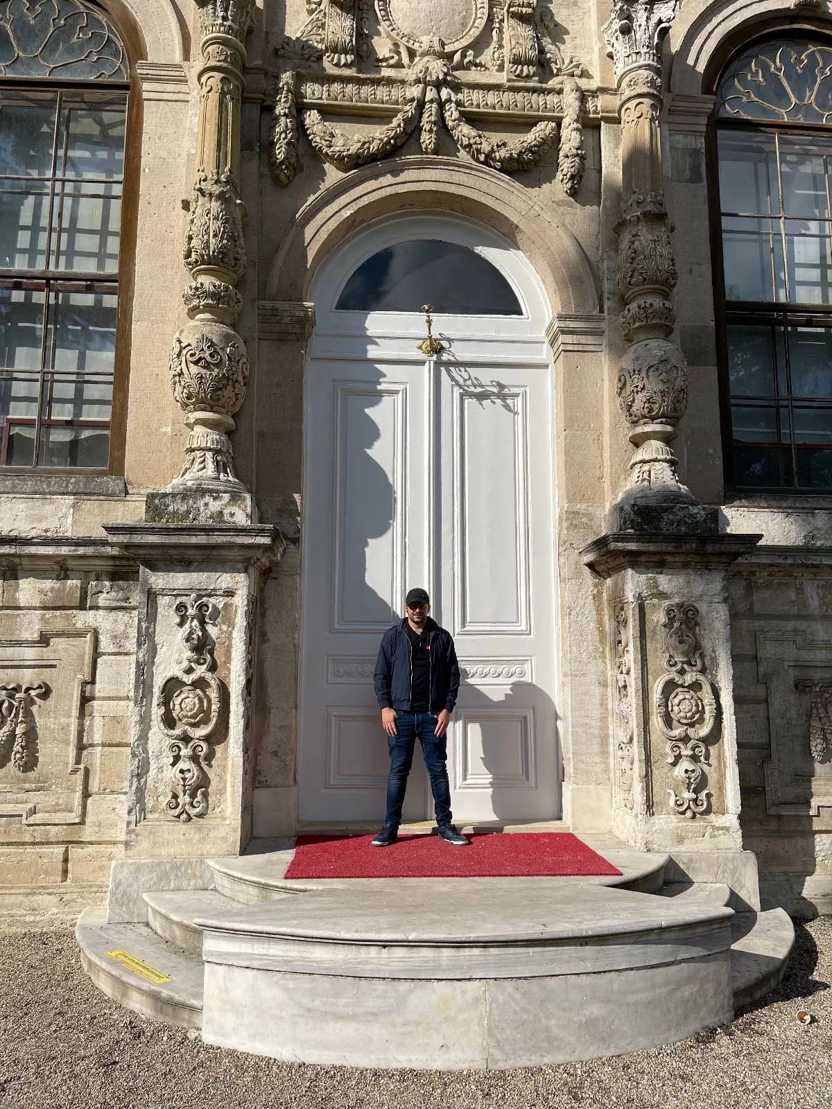 A person stands on a red carpet in front of an ornate, tall white door framed by detailed stone carvings and two large windows, with sunlight casting shadows on the building facade.