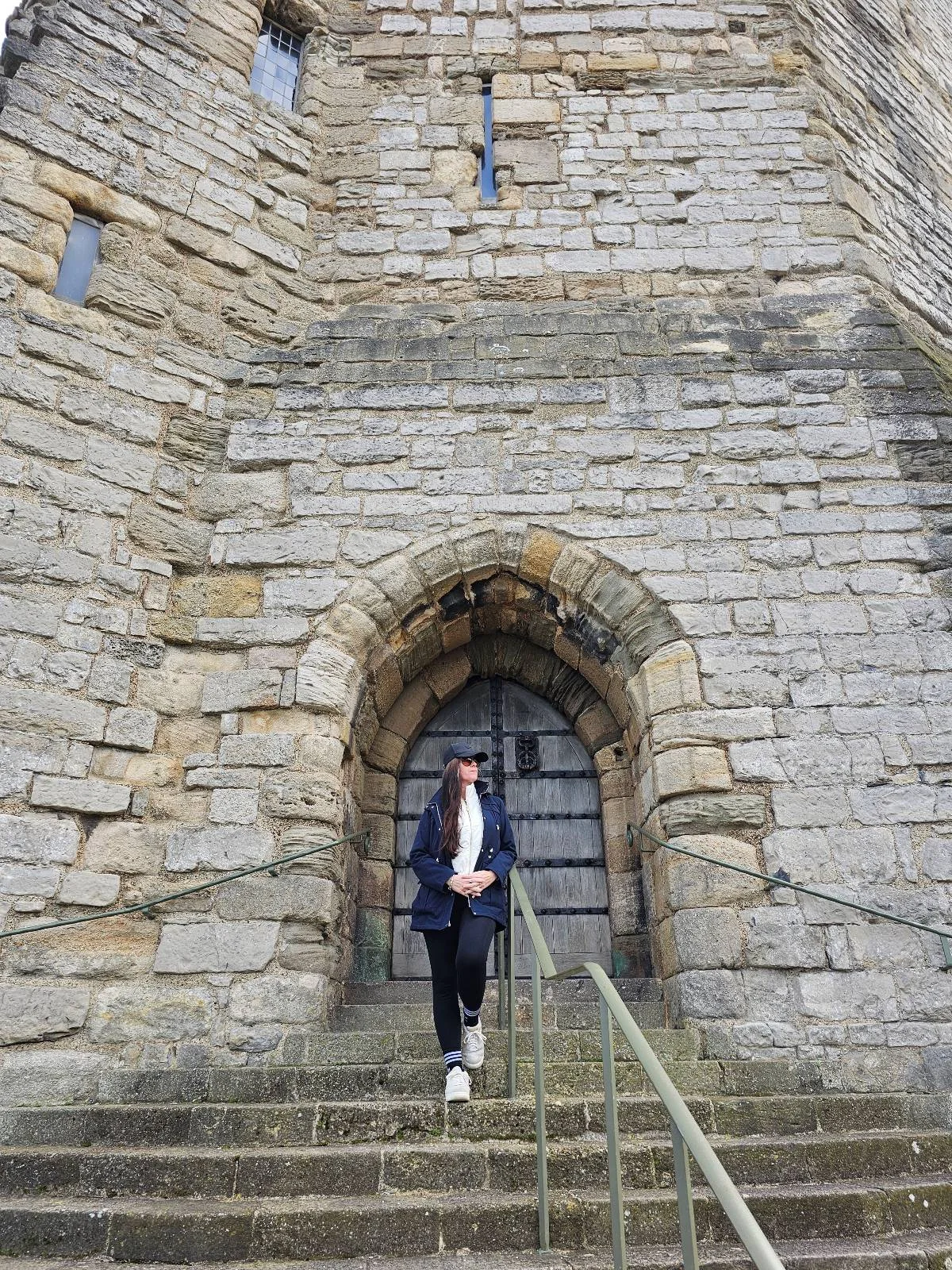 A person in a dark jacket and white shoes walks down stone steps in front of an arched doorway, set in the wall of an old stone building or castle.
