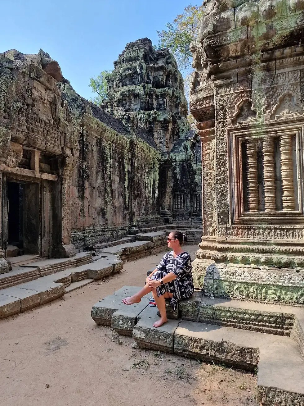 A person in a patterned dress sits on ancient stone ruins, enjoying the sunlight. The background features ornate carvings and blue sky.